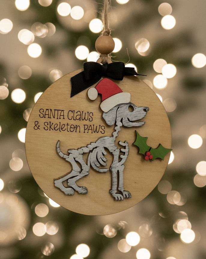 Wooden Christmas ornament with skeleton dog design and text 'Santa Claus & Skeleton Paws' against a blurred Christmas tree background.